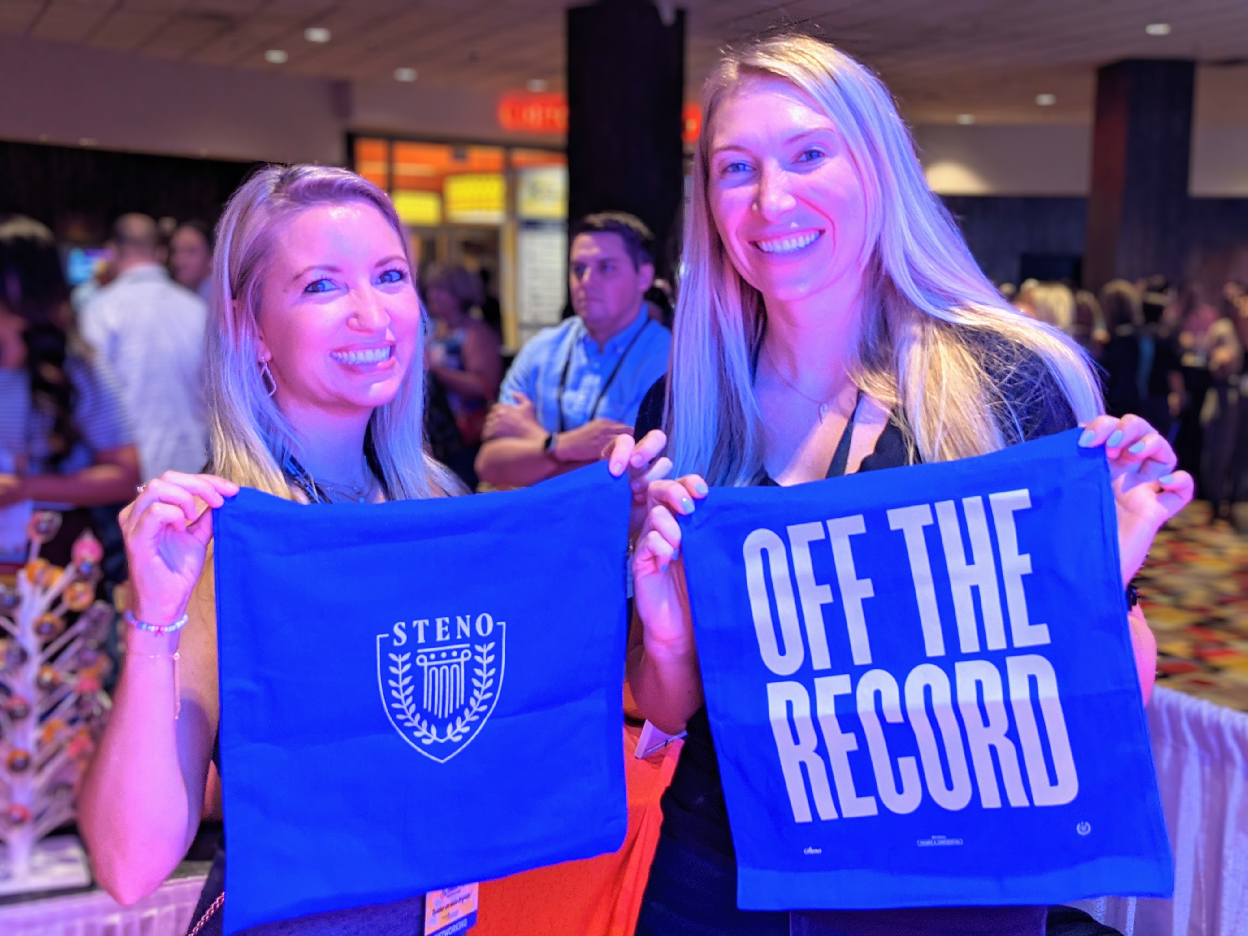 Two women hold blue tote bags that display a Steno logo and "Off the Record" 
