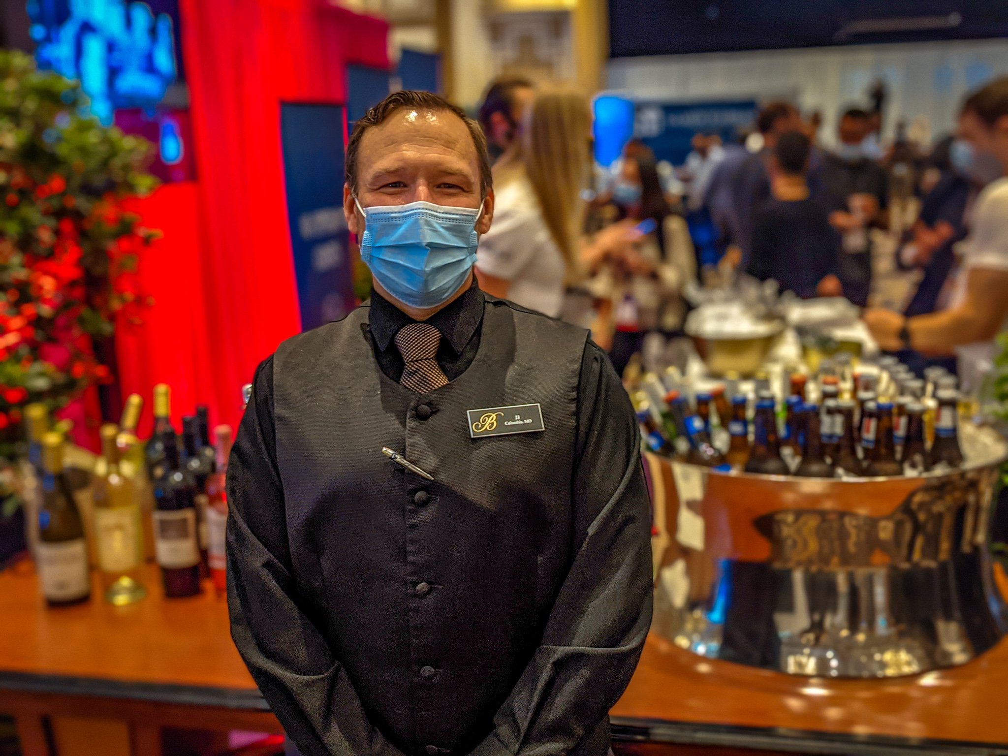 A man wearing a nametag that says "JJ" stands in front of a bar.